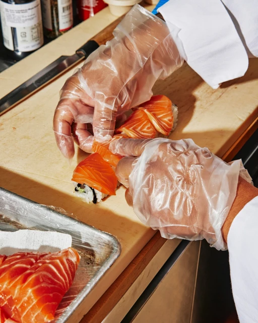 Close-up shot of a chef wearing clear plastic gloves preparing fresh salmon sushi rolls on a wooden cutting board in a kitchen.