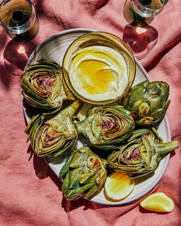 Overhead food photography of artichokes and dipping sauce on a plate, placed on a pink linen tablecloth with natural window light.