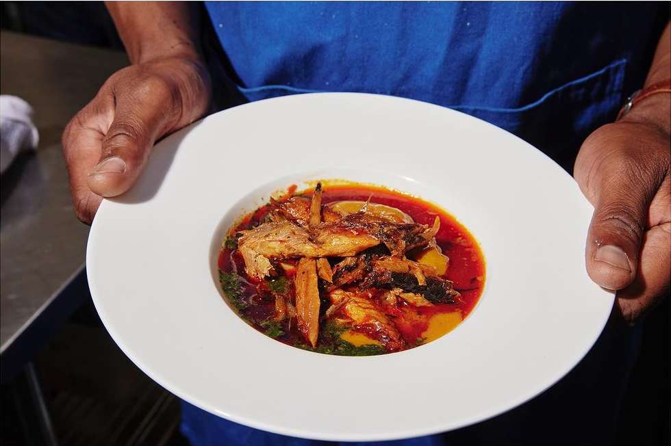 Close-up of dark-skinned hands holding a bowl of fish stew in red broth, with green garnish and potatoes, on a white plate.