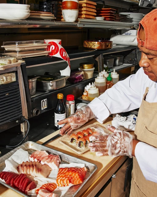Sushi chef in an indoor restaurant kitchen prepares sushi rolls and sashimi with raw fish on a metal tray, wearing plastic gloves.