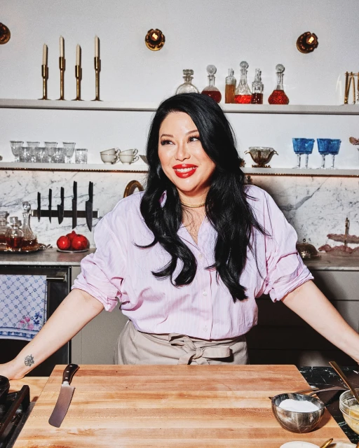 Woman in kitchen preparing food on wooden cutting board. Adult female with black hair wearing pink shirt and apron, countertop setting.