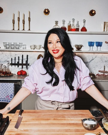 Woman in kitchen preparing food on wooden cutting board. Adult female with black hair wearing pink shirt and apron, countertop setting.