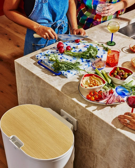 Two people preparing food on a marble countertop with hummus, vegetables, drinks, and a white waste bin in a home kitchen.