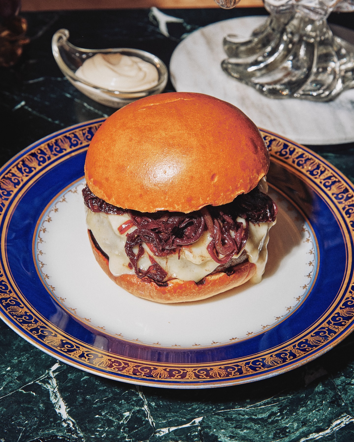 Gourmet hamburger on a white plate with a blue and gold rim, featuring melted cheese, red onions, and a brioche bun, served on marble.