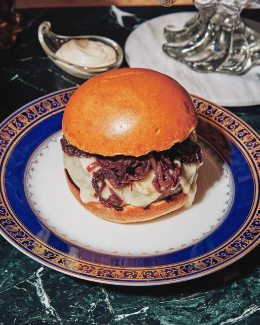Gourmet hamburger on a white plate with a blue and gold rim, featuring melted cheese, red onions, and a brioche bun, served on marble.