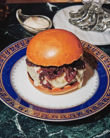 Gourmet hamburger on a white plate with a blue and gold rim, featuring melted cheese, red onions, and a brioche bun, served on marble.