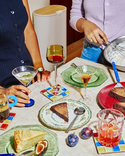 Table featuring slices of cake, cocktails, fruit, and plates. Two people are partially visible at the table with drinks and food.