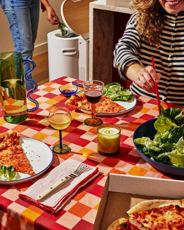 Two people share pizza and salad at a table covered with a patterned tablecloth. Food scraps are discarded in a compost bin.