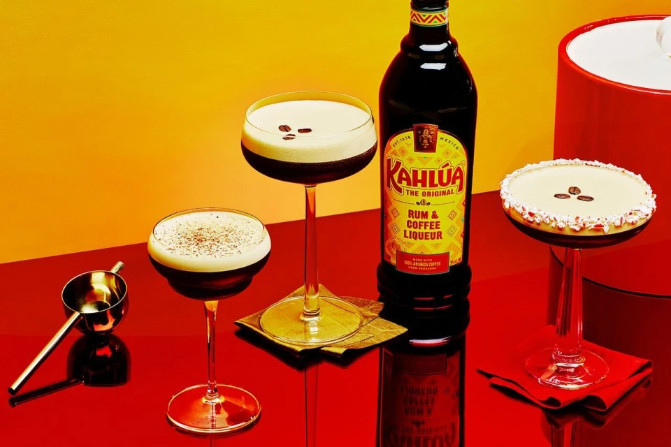 Three espresso martini cocktails with coffee beans, Kahlua bottle, and a gold bar spoon against a red and yellow gradient backdrop.