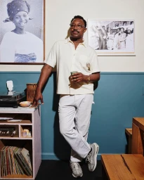 Man wearing a cream button-up shirt and white pants leaning against a turntable stand in a teal and white room photographed by Laura Murray.