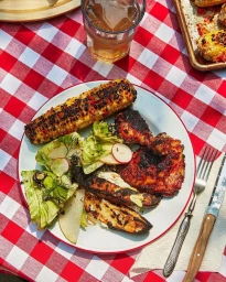 Overhead food photography of a ceramic plate with grilled chicken, corn on the cob, and salad on a red and white checkered tablecloth.