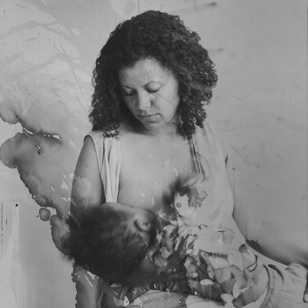 Black and white portrait of an adult woman breastfeeding a baby in a close-up studio setting with a textured background.