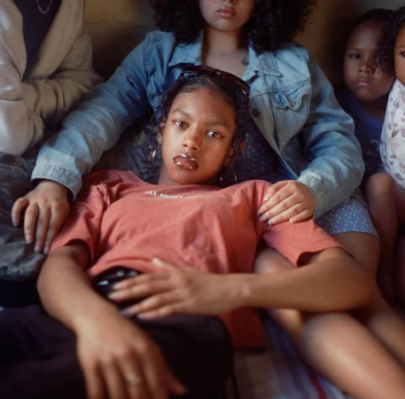A group of African American women and children are indoors, with one woman laying on the others lap while facing the camera.