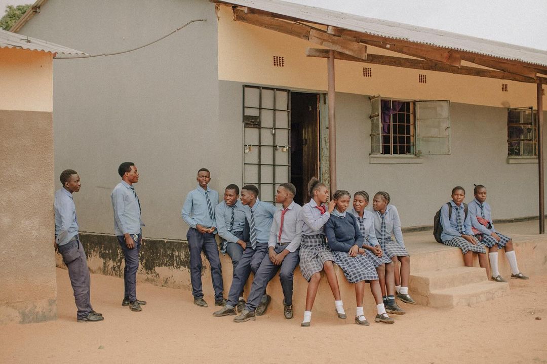 Group of African students in school uniforms gather in front of a building with open windows, exhibiting a mix of relaxed and engaged postures.