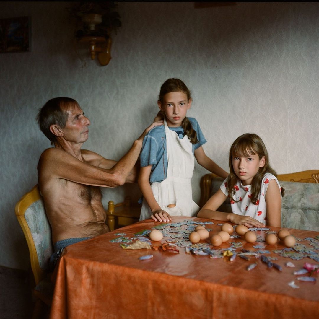 An elderly man stands shirtless with his hands on a young girl's shoulders, with another young girl seated at a table with a puzzle and eggs.