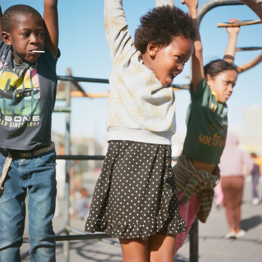 Three children hang from a metal bar structure on a sunny day. Two girls and a boy are outside; one child has a lollipop.