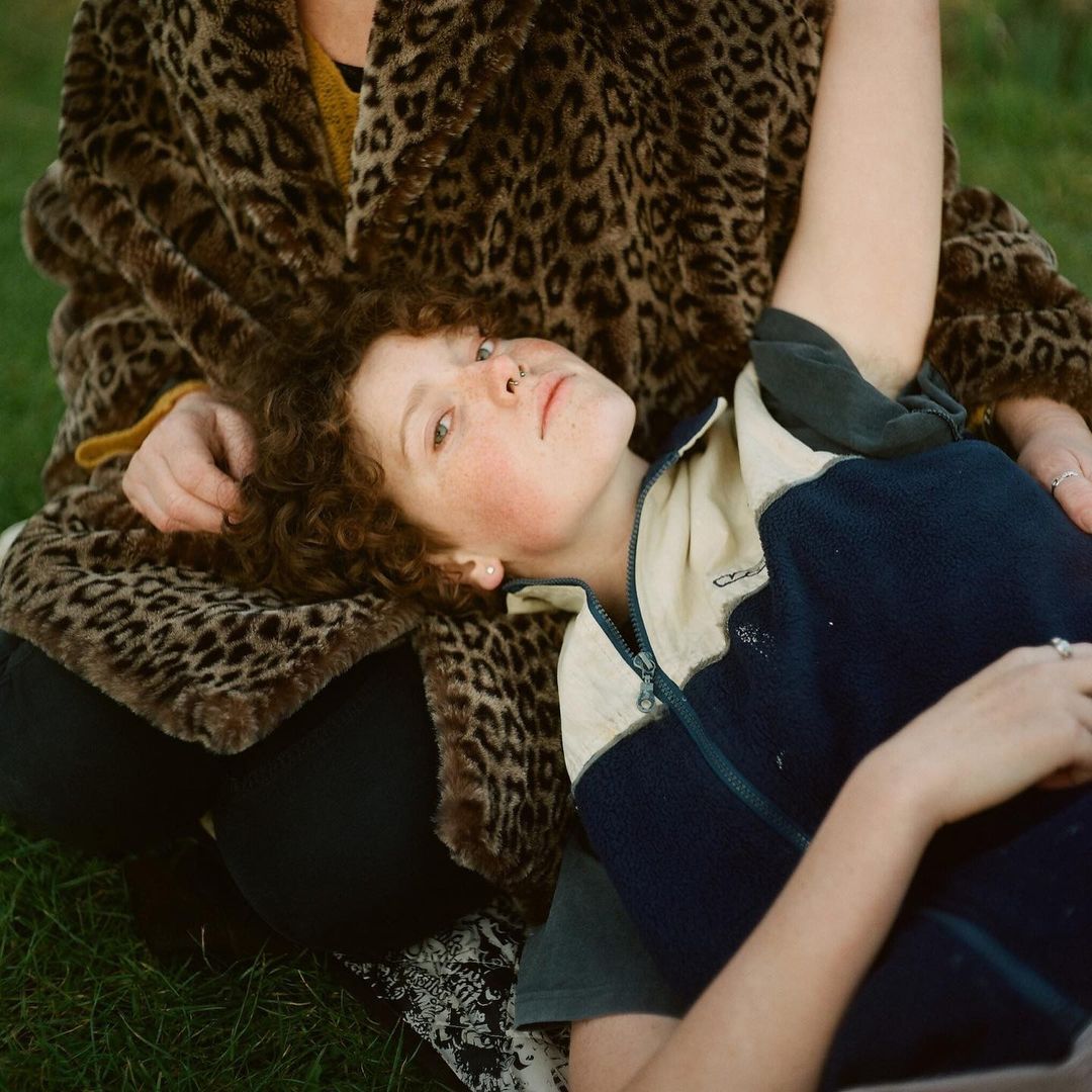 A child with curly red hair lying down with their head on a person wearing a brown leopard print jacket in a grassy outdoor setting.