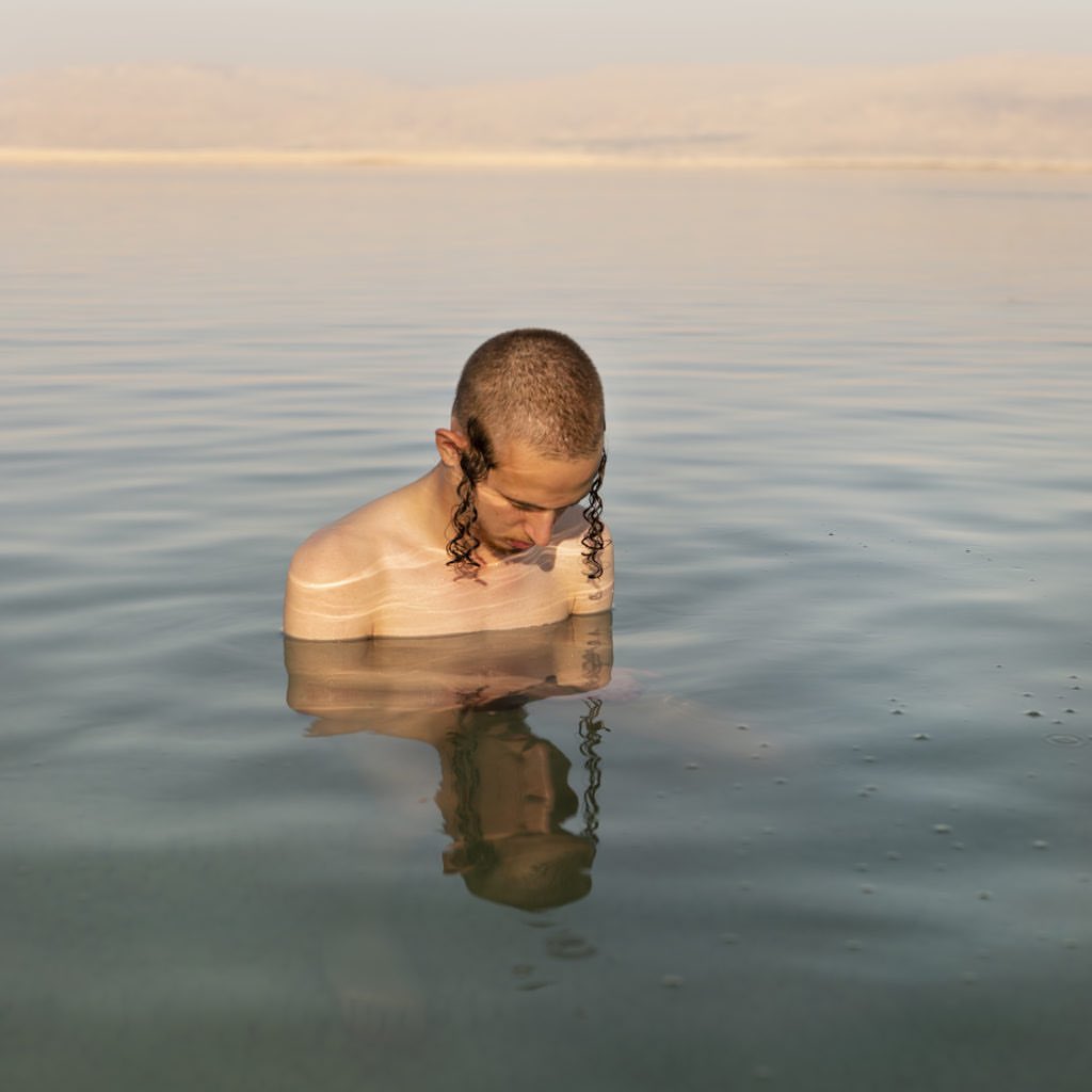 A man with hair at his temples stands chest-deep in water during the daytime. The horizon is visible in the background. 