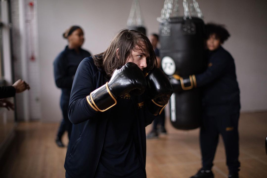 A young adult boxer with long brown hair wearing gloves and a blue shirt in a gym setting, looking focused during a training session.