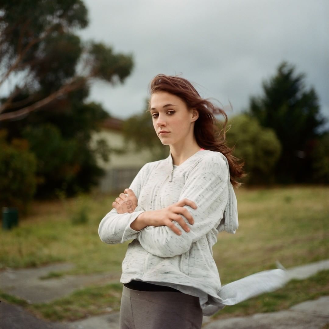 A young woman with red hair, wearing a gray hoodie and dark pants, stands with crossed arms in an outdoor suburban setting under cloudy skies.
