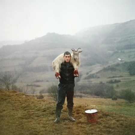 Young man carrying a white goat across his shoulders standing on a grassy hill with misty mountain landscape behind him by Laura Pannack