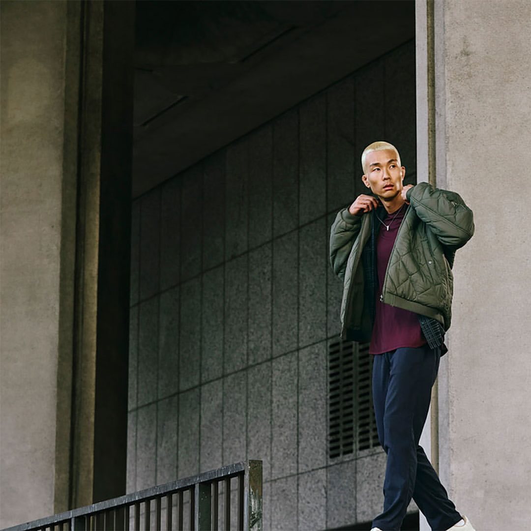 Man in a quilted jacket stands outside a concrete building with a rail, looking over his shoulder in a fashion style shot.