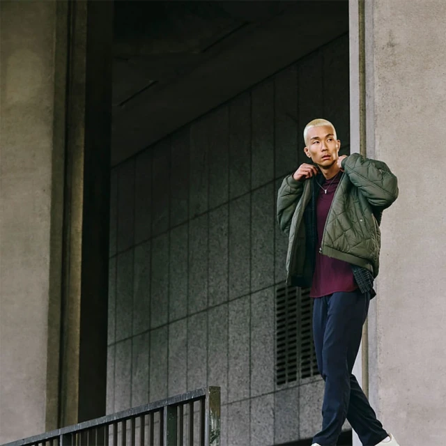 Man in a quilted jacket stands outside a concrete building with a rail, looking over his shoulder in a fashion style shot.