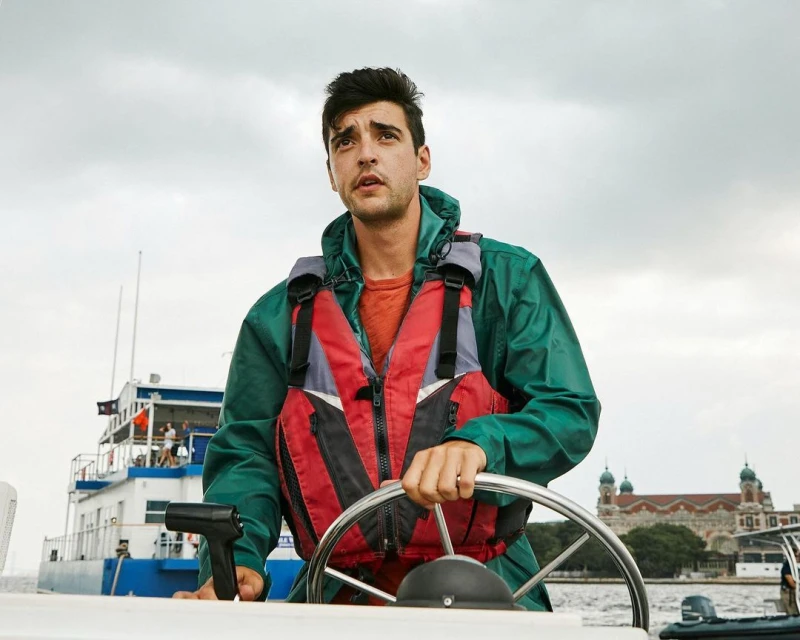 Man in life vest steers boat on water under cloudy sky, with building visible in the background. Safety and water transportation.