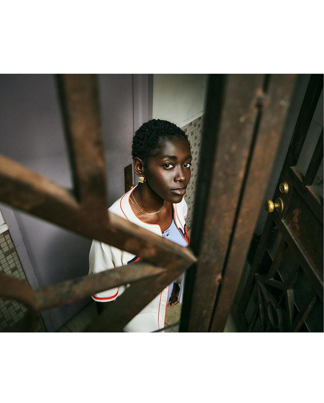 Low-angle shot of a stylish young Black woman with short hair looking up in a stairwell, wearing a white baseball jacket and gold earrings.