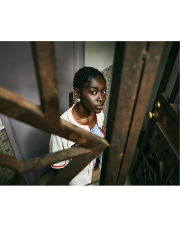 Low-angle shot of a stylish young Black woman with short hair looking up in a stairwell, wearing a white baseball jacket and gold earrings.