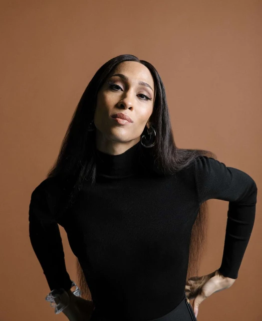 Portrait of an adult woman with long, straight dark hair wearing a black turtleneck, posing with hands on her hips against a tan background.