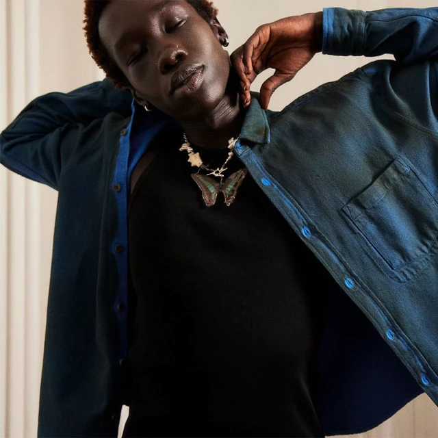 Close-up studio shot of a dark-skinned model wearing a black top, blue button-down shirt, and statement butterfly necklace posing.
