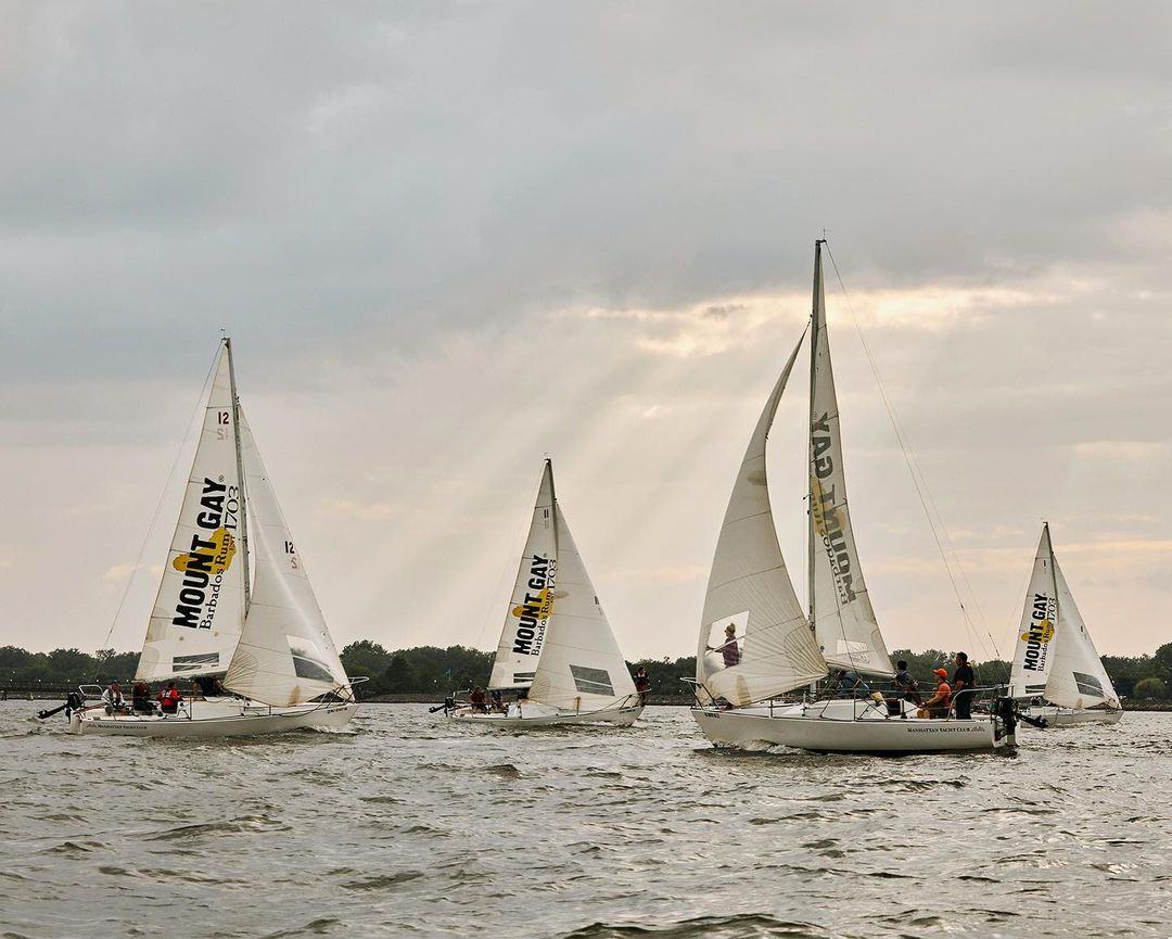 Multiple sailboats with white sails and crews sailing on the water under a cloudy sky with sun rays breaking through.