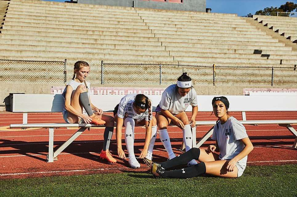 Four female athletes in sports attire sit on a bench at a stadium track, tying their shoelaces and socks in bright sunlight.