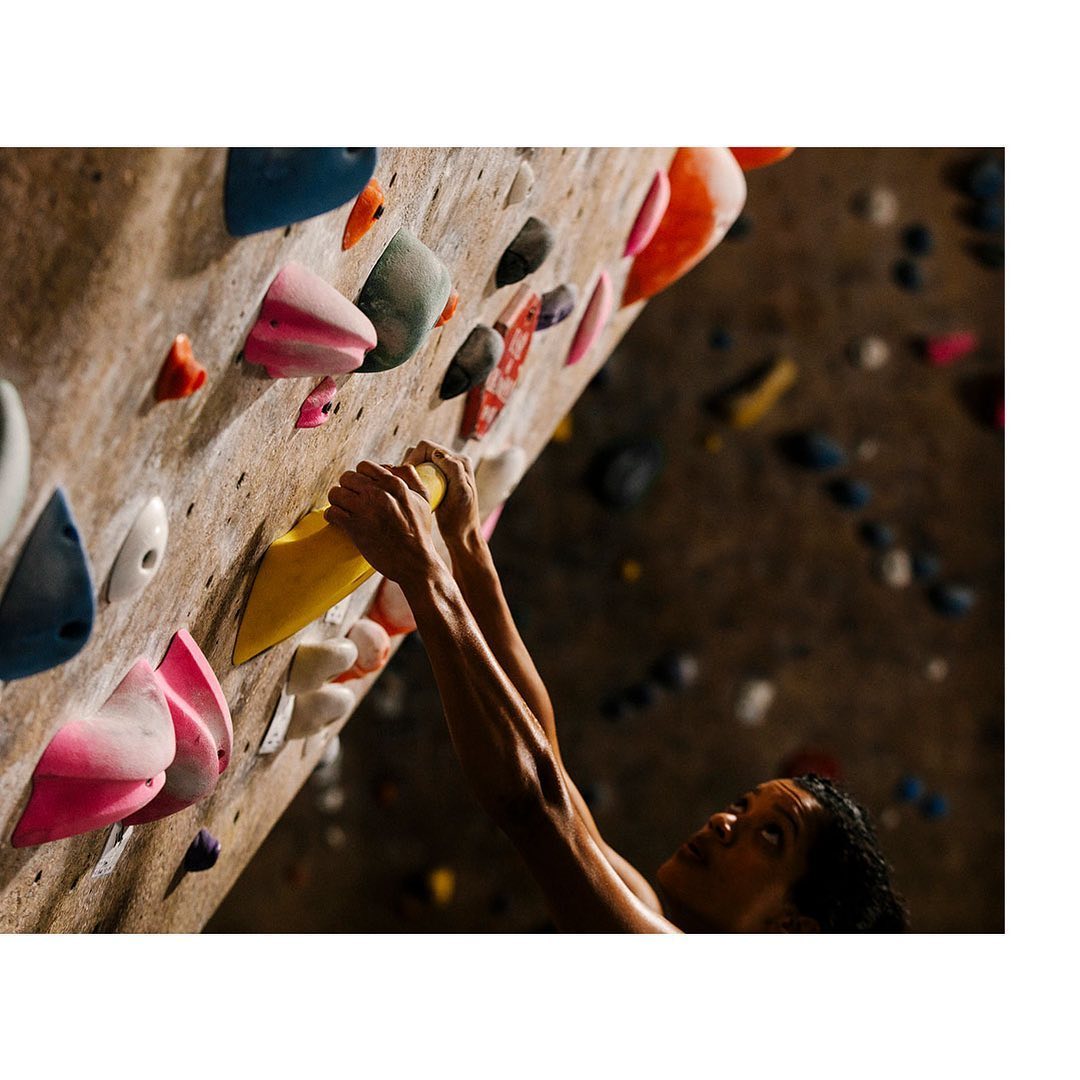 Adult male rock climber ascending a climbing wall indoors, holding onto yellow climbing hold with various shaped holds on wall.