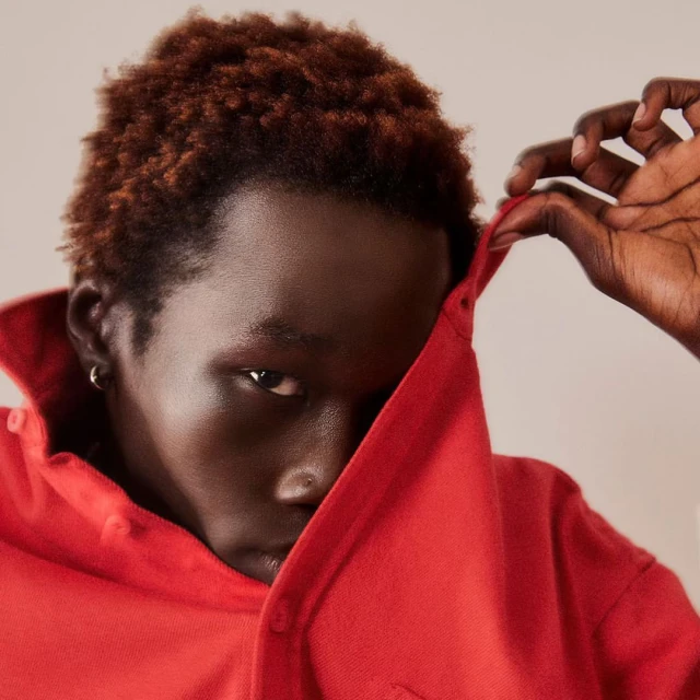 Young man with brown hair in a red button-down shirt partially covering his face in a studio portrait with an intense gaze.