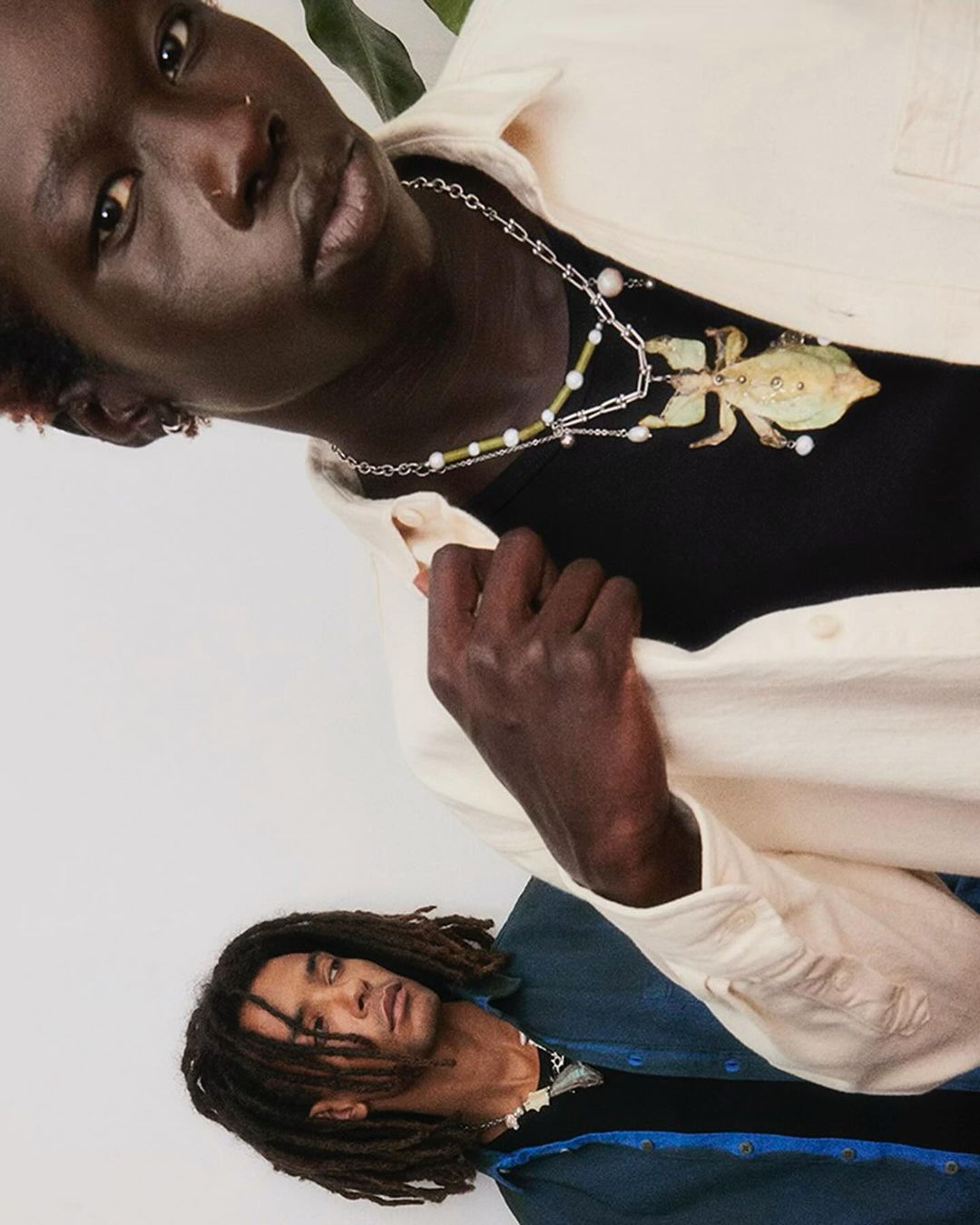 Two dark-skinned adult men wear unique jewelry in a studio setting, captured in a medium shot with soft lighting and neutral tones.