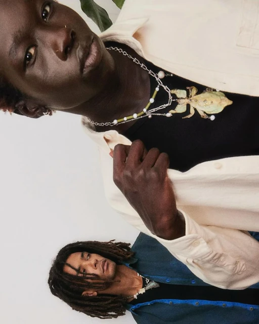 Two dark-skinned adult men wear unique jewelry in a studio setting, captured in a medium shot with soft lighting and neutral tones.