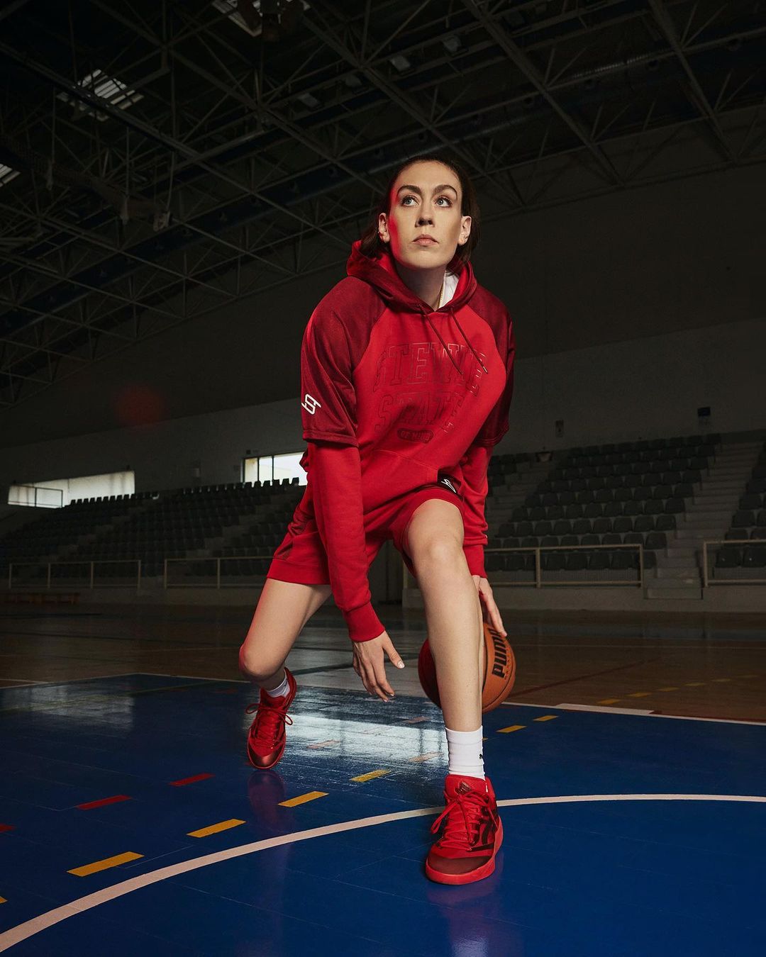 Female athlete in red activewear dribbles a basketball on an indoor court, illuminated by stadium lights.