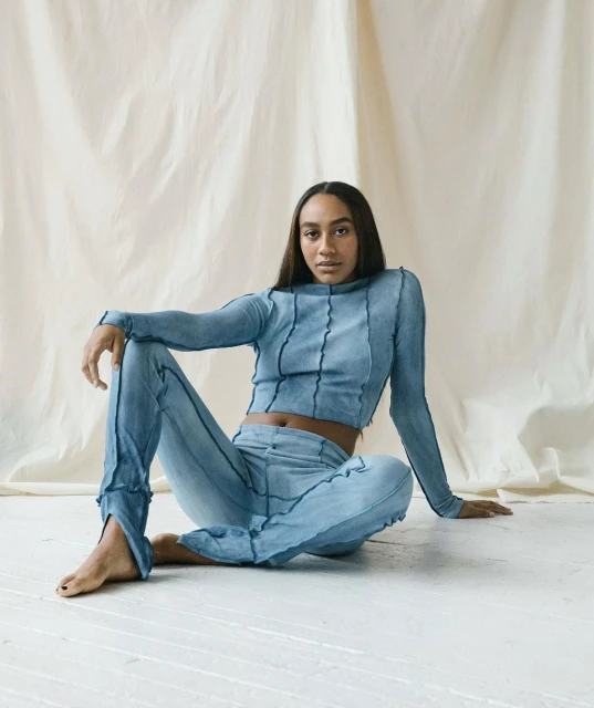 Woman poses in light blue, long-sleeved crop top and matching pants, seated on the floor, with a cream-colored fabric backdrop.