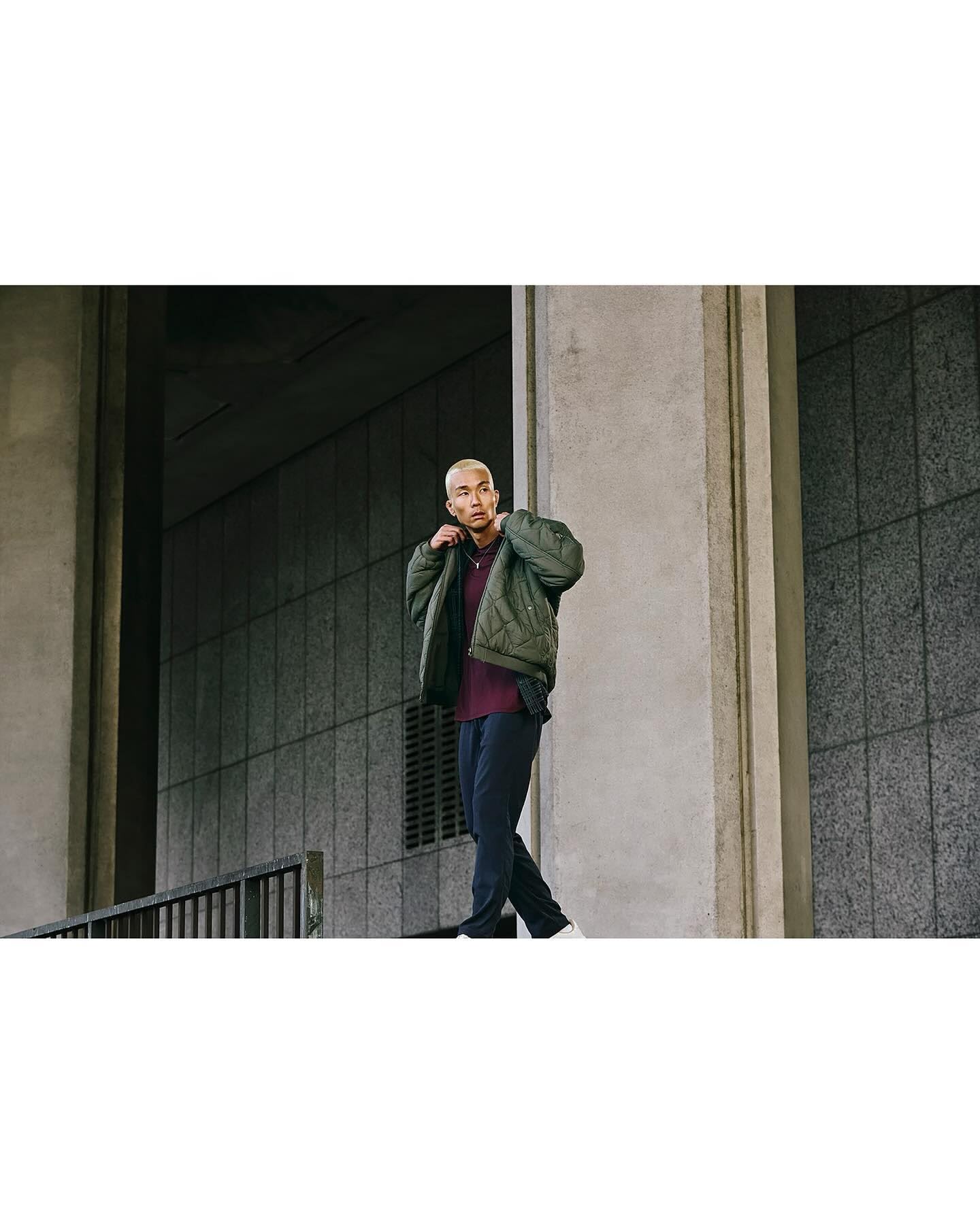 A stylish man in a green jacket walks outdoors against a concrete wall. He is wearing dark pants and light sneakers.
