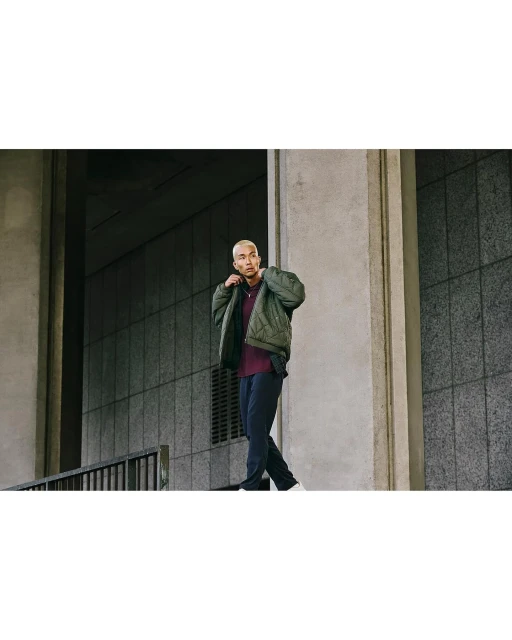 A stylish man in a green jacket walks outdoors against a concrete wall. He is wearing dark pants and light sneakers.