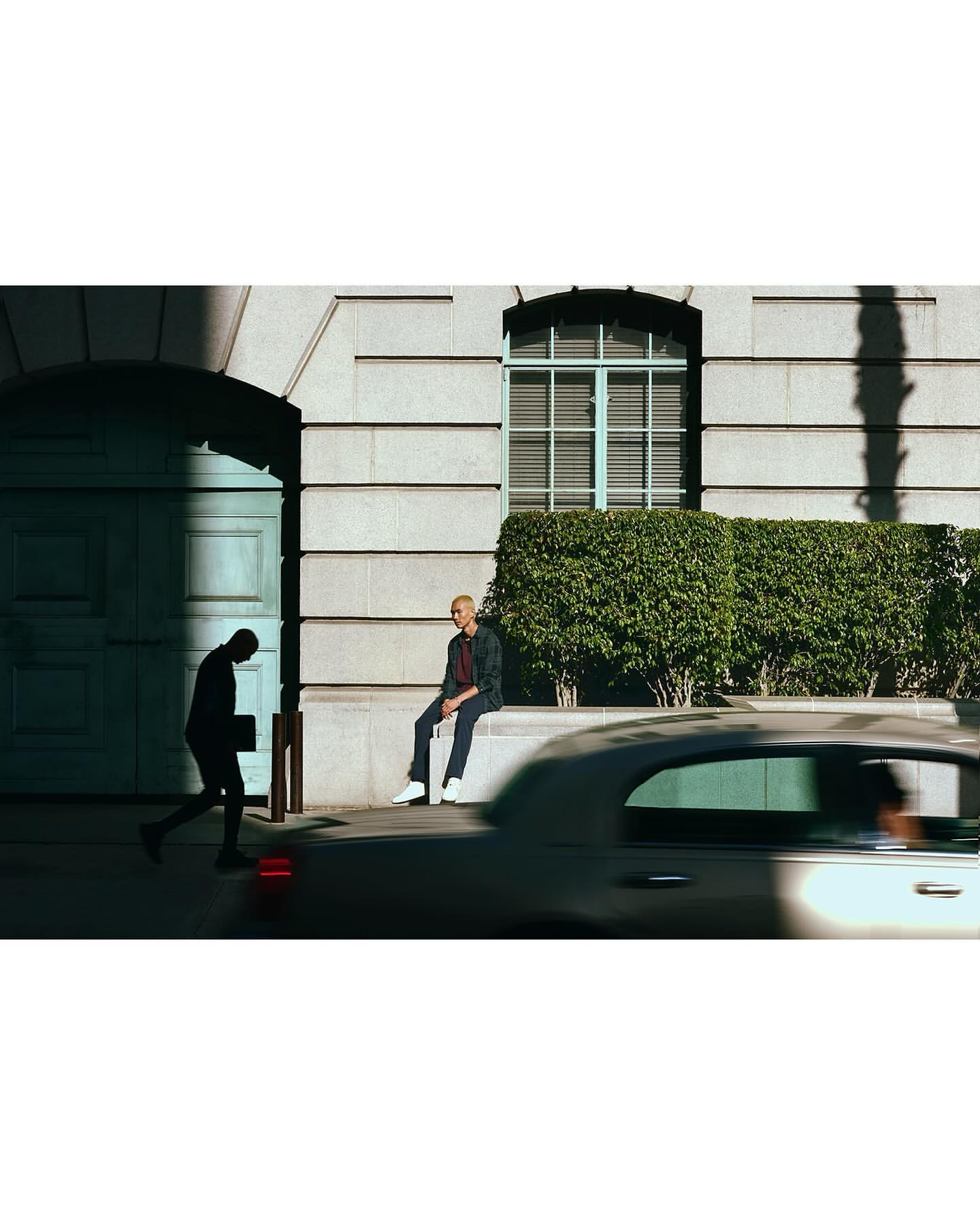 Man in jacket sitting on a ledge in an urban scene while another man walks by an archway, a car passes, and sunlight streams in.