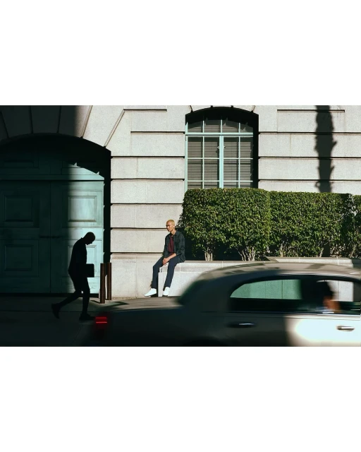 Man in jacket sitting on a ledge in an urban scene while another man walks by an archway, a car passes, and sunlight streams in.
