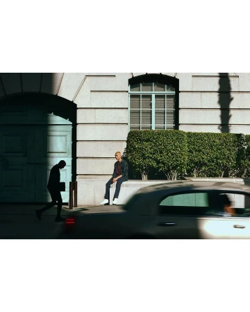 Man in jacket sitting on a ledge in an urban scene while another man walks by an archway, a car passes, and sunlight streams in.