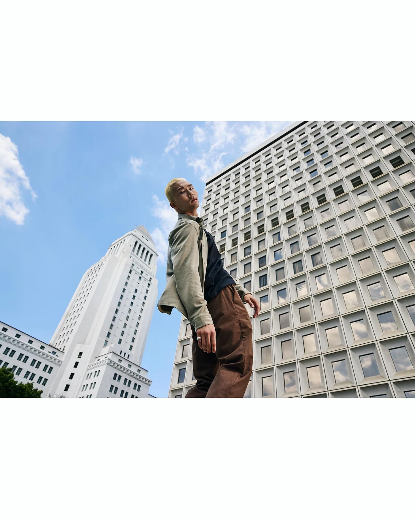 Low-angle shot of a young man with blonde hair standing against tall modern and classical buildings under a blue sky.