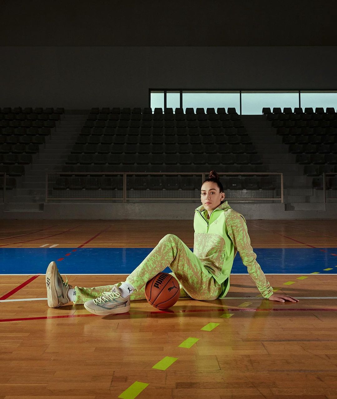 A woman in athleisure wear sits on a basketball court with a ball, posing for a fashion or sports editorial photograph indoors.