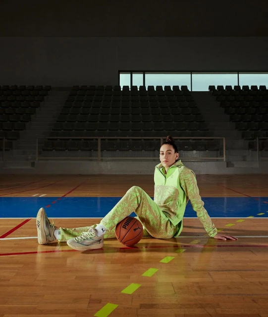 A woman in athleisure wear sits on a basketball court with a ball, posing for a fashion or sports editorial photograph indoors.