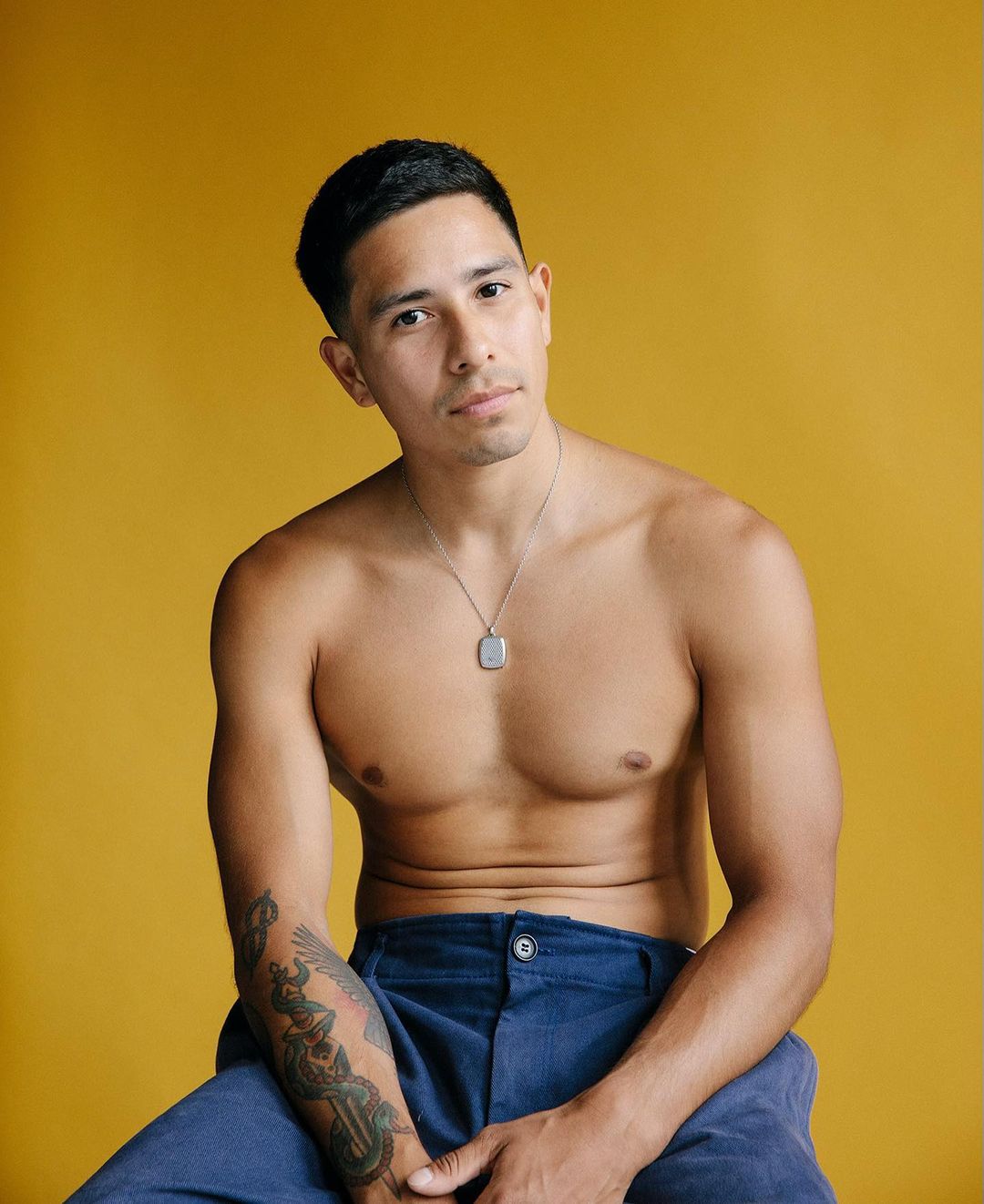 Shirtless man with arm tattoos wearing blue trousers wearing a silver pendant necklace against a yellow backdrop