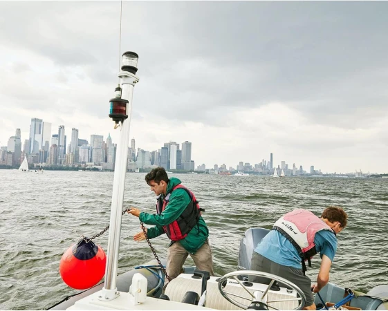 Two people in life vests on a small boat on the water with a cityscape in the background under an overcast sky.